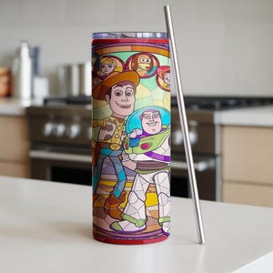 Puede incluir: Un vaso de acero inoxidable con un dise&ntilde;o de vidriera con los personajes Woody y Buzz Lightyear de Toy Story. El vaso tiene una tapa y base rojas, y una pajita plateada. El fondo es una cocina.
