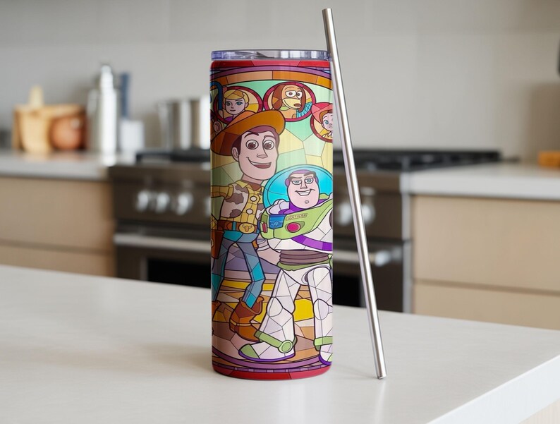 Puede incluir: Un vaso de acero inoxidable con un dise&ntilde;o de vidriera con los personajes Woody y Buzz Lightyear de Toy Story. El vaso tiene una tapa y base rojas, y una pajita plateada. El fondo es una cocina.