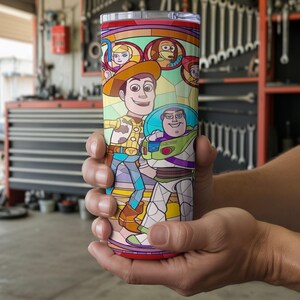 Puede incluir: Un vaso de acero inoxidable con un colorido dise&ntilde;o de vidriera que presenta a los personajes Woody y Buzz Lightyear de la pel&iacute;cula animada Toy Story. El vaso tiene un borde rojo y una tapa transparente. El dise&ntilde;o incluye otros personajes de la pel&iacute;cula.