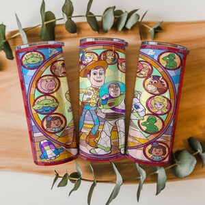 Puede incluir: Tres vasos de acero inoxidable con un colorido dise&ntilde;o de vidrieras que presenta personajes de una popular pel&iacute;cula de animaci&oacute;n. Los vasos tienen un borde y una base rojos, y una tapa transparente. El dise&ntilde;o incluye a Woody, Buzz Lightyear y otros personajes.