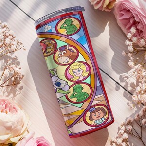 Puede incluir: Un vaso de acero inoxidable con un dise&ntilde;o de vidriera que presenta personajes de la pel&iacute;cula animada Toy Story. El vaso tiene un borde rojo y una tapa transparente. El dise&ntilde;o incluye a Woody, Buzz Lightyear y otros personajes.
