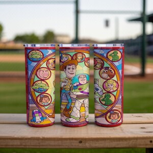 Puede incluir: Tres vasos aislados rojos y plateados con un dise&ntilde;o de vidriera que presenta personajes de la pel&iacute;cula animada Toy Story. Los vasos son cil&iacute;ndricos y est&aacute;n sobre una superficie de madera.