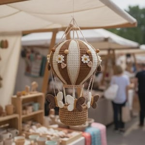 Puede incluir: Un globo aerostático de ganchillo hecho a mano en colores marrón y crema, adornado con motivos de margaritas y detalles de mariposas. El globo está suspendido, mostrando una cesta debajo, perfecto para la decoración de la guardería o un toque caprichoso.