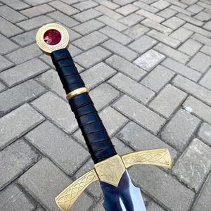 May include: A sword featuring a black leather-wrapped grip, a gold-colored crossguard with intricate etched patterns, and a circular pommel set with a faceted red gemstone. The blade is metallic silver with a polished finish and a central fuller.