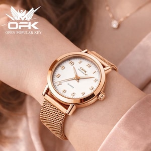 Peut inclure: Montre-bracelet en or rose avec un cadran blanc, des chiffres et des aiguilles noirs. La montre a un bracelet en maille et un cadran rond. La marque "ORK" est visible sur le cadran. Mouvement à quartz.