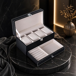 Peut inclure: Un coffret à montres en cuir noir avec des coutures blanches et un fermoir argenté. La section supérieure comporte quatre coussins pour montres, et le tiroir inférieur des emplacements pour bagues et un compartiment. L'intérieur est beige clair.