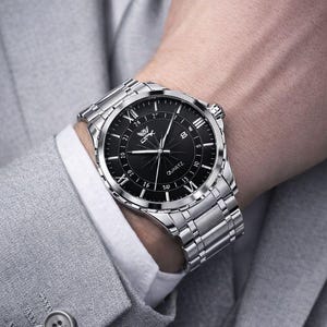Peut inclure: Montre à quartz OPK argentée avec un cadran noir, des chiffres romains et un affichage de la date. La montre est sur un bracelet en argent. La montre est portée avec un costume gris et une chemise blanche.