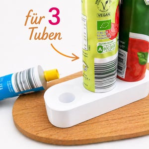 Könnte beinhalten: Ein weißer Kunststoff-Tubenhalter mit zwei Tuben Lebensmittel und einer Tube auf einer Holzoberfläche. Der Halter hat zwei Löcher für die Tuben. Der Text "für 3 Tuben" ist im Bild sichtbar.