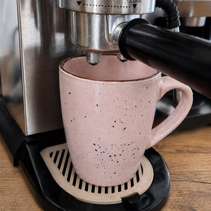 Könnte beinhalten: Eine hellrosa Keramiktasse mit dunkelbraunen Sprenkeln unter dem Siebträger einer Espressomaschine aus Edelstahl. Die Maschine hat einen schwarzen Griff und einen silbernen Brühkopf mit dem aufgedruckten Text INSERT und CLOSE.