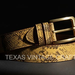 May include: A snakeskin-patterned belt with a gold-colored buckle. The belt is yellow and brown with a textured surface. The text "TEXAS VINTAGE CRAFT" is printed on the belt.