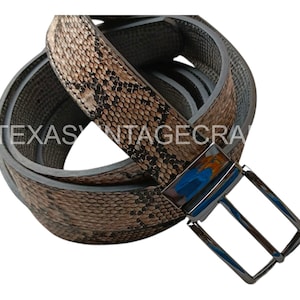 Op de afbeelding: Een opgerolde riem met een slangenpatroon in bruin- en beigetinten. De riem heeft een zilverkleurige gesp. De tekst "TEXAS VINTAGECRA" is zichtbaar op de riem.