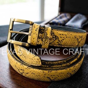 May include: A yellow snakeskin-patterned belt with a gold-colored buckle. The belt is coiled on a wooden surface. The text "TEXAS VINTAGE CRAFT" is visible on the belt. A watch and accessories are in the background.