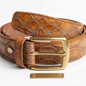 Könnte beinhalten: Ein brauner Ledergürtel mit einem strukturierten, schuppenartigen Muster. Der Gürtel hat eine goldfarbene Schnalle und ein kleines rechteckiges Etikett mit der Aufschrift "TEXAS VINTAGE CRAFT". Der Gürtel ist aufgerollt und zeigt sein Design.