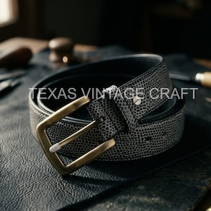 Puede incluir: Un cinturón de cuero negro con un patrón moteado y una hebilla dorada. El cinturón está enrollado sobre una superficie oscura, con el texto "TEXAS VINTAGE CRAFT" visible en el cinturón.