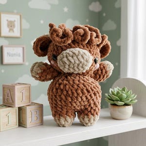May include: A brown and beige crocheted plush toy shaped like a Highland cow with a textured, fuzzy yarn finish, dark round eyes, and small pink cheeks. The stuffed animal features curly hair on top of its head and a light-colored muzzle.