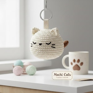 May include: A round, cream-colored crochet cat keychain featuring a small brown tail, embroidered black eyes, and whiskers. A metal ring is attached to the top. A rectangular label displays the text Mochi Cats Handmade Crochet Art.