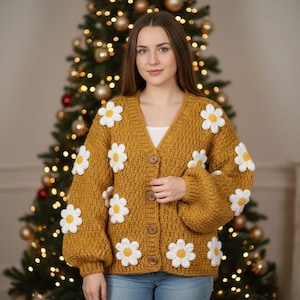 Puede incluir: Un cárdigan de manga larga color amarillo mostaza con cuello en V y cuatro botones marrones en la parte delantera. La prenda de punto está decorada con varias margaritas blancas grandes de ganchillo con centros amarillos en el frente y mangas.