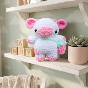 May include: A plush cow toy made of white and pink yarn with a soft, fuzzy texture. The cow features black eyes, a pink snout, and pink patches on its ears, feet, and body. It wears a light green crossbody bag with a small pink strawberry charm.