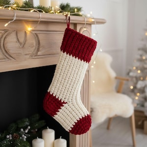 Könnte beinhalten: Ein grob gestrickter Weihnachtsstrumpf mit gerippter Textur, einem dunkelroten Umschlag, einem cremefarbenen Körper sowie einer passenden roten Ferse und Spitze. Der Strumpf hängt an einer Schlaufe aus Stoff an einem hölzernen Kaminsims.