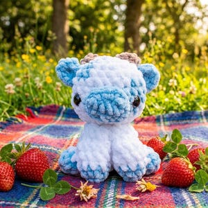 May include: A small, plush, crocheted cow toy featuring a soft, white and light blue mottled yarn texture, two small brown horns, and large, round, black plastic eyes, sitting upright with a rounded head and four short, stubby legs.