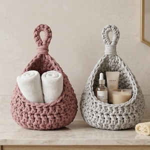 May include: Two handmade crochet hanging baskets, one in a dusty rose and one in grey, with looped handles. The pink basket holds rolled white towels, the grey basket contains skincare products and a candle. Bathroom decor.