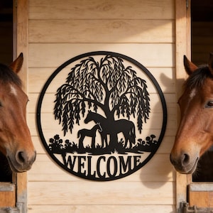 May include: A black metal welcome sign with a tree and horse silhouette is centered on a wooden wall. Two brown horses are in stalls on either side of the sign. The word "WELCOME" is at the bottom of the sign.