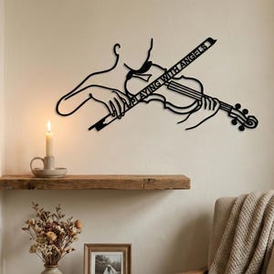 Puede incluir: Arte mural de metal negro que representa a una persona tocando el violín. El texto "PLAYING WITH ANGELS" está en el violín. Montado en una pared blanca sobre una estantería de madera. Una vela encendida y flores secas están en la estantería.