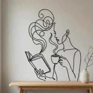 以下が含まれることがあります： 本を読み、コーヒーカップを持っている女性を描いた黒い金属製の壁の芸術。 このアートワークは、女性の髪が上向きに流れる連続線画スタイルです。 小さな白い花瓶と枝が木製のテーブルの上にあります。
