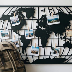 Pode incluir: Decoração de parede de mapa-múndi em metal preto com uma grelha, exibindo fotos presas com prendedores de roupa de madeira. Uma mochila de lona está em primeiro plano. As fotos mostram destinos de viagem. O mapa tem cerca de 70 cm de largura.