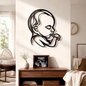 Op de afbeelding: Een zwart metalen wanddecoratie met een doorlopende lijntekening van een babyprofiel. Het ontwerp toont het hoofd en bovenlichaam van een zuigeling met een hand bij de mond, uitgevoerd in een vloeiende en onderling verbonden stijl.