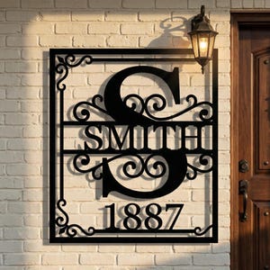 Puede incluir: Letrero de nombre familiar de metal negro con la letra "S", el nombre "SMITH" y el año "1887". El letrero tiene adornos decorativos y está montado en una pared de ladrillo blanco. Una lámpara de exterior de estilo vintage está montada encima.
