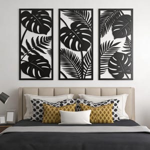 Könnte beinhalten: Drei schwarz gerahmte Wandkunst-Paneele mit tropischen Blattmotiven, darunter Monstera- und Farnblätter. Das Kunstwerk hängt über einem Bett mit beigem Kopfteil, Kissen und einer dunkelgrauen Tagesdecke. Eine Lampe und Bilderrahmen stehen auf dem Nachttisch.