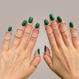 May include: Two hands with green nail polish are adorned with multiple silver finger rings. The rings are thin and minimalist in design, creating a modern and stylish look. The rings are worn on various parts of the fingers.