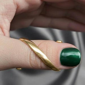 May include: A gold-colored ring wraps around a finger, with a small gold accent on the nail. The nail is painted with a dark green polish. The ring has a smooth, curved design.