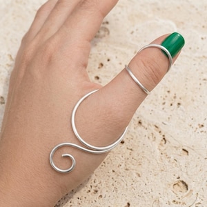 May include: A silver-colored finger ring with a unique, swirling design. The ring wraps around the thumb and finger, featuring a curved shape and a spiral detail. The thumb has a green nail polish.