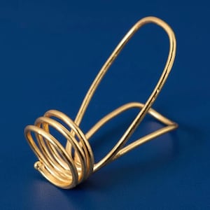 以下が含まれることがあります： ユニークなデザインのゴールドカラーの金属製ナプキンホルダー。ホルダーは、コイル状のベースとアーチ型のトップが特徴で、エレガントでモダンな外観を作り出しています。ナプキンホルダーは、光沢のある反射性の金属で作られています。