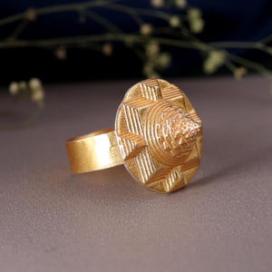 Pode incluir: Um anel dourado com um design circular texturizado e um centro em forma de cone. A banda do anel é de ouro liso. O anel é provavelmente feito de metal e é exibido numa superfície neutra.