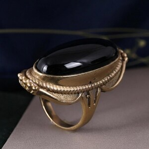 May include: A vintage-style ring featuring a large, oval-shaped black stone set in a gold-toned metal. The ring has an ornate, textured design around the stone and on the band, showcasing a classic, antique aesthetic. The ring is a statement piece.
