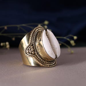 May include: A brass ring featuring a large, white seashell. The ring has an ornate design with braided and floral details. The seashell is prominently displayed, creating a unique and eye-catching piece of jewelry.