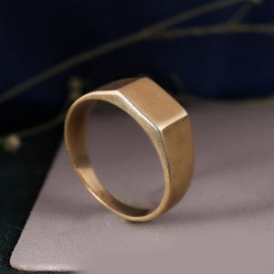 Puede incluir: Un anillo de sello en tono dorado con una superficie superior rectangular plana y lisa, y una banda sólida sin adornos. El metal tiene un acabado mate y cepillado, mostrando un diseño simple y limpio sin grabados ni elementos decorativos.