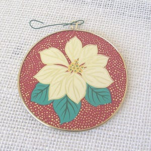 May include: A circular ornament featuring a cream-colored poinsettia flower with green leaves centered on a deep red background with gold-colored speckles. The design is enclosed in a thin gold-colored metal frame with a green wire hook at the top.