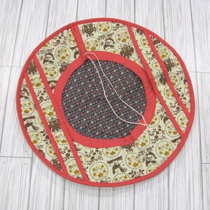 May include: A circular fabric tree skirt featuring a cream-colored outer ring with a brown and yellow floral and woodland animal pattern, bordered by red trim. The center is a dark blue fabric with a small red and white floral print and white ties.