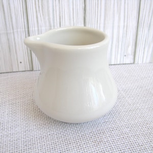 May include: A small, off-white ceramic creamer pitcher with a rounded body and a pouring spout. The creamer is sitting on a white textured surface, with a white wooden background.