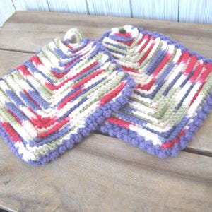 May include: Two handmade, square crochet potholders. Each potholder features a chevron pattern in red, white, green, and blue yarn, with a purple border and hanging loop. The potholders are displayed on a wooden surface.