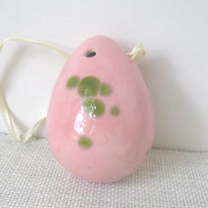 May include: A pink ceramic pendant with a glossy finish, featuring irregular green spots. The pendant has a small hole at the top and is strung with a cream-colored ribbon. The egg-shaped ornament is displayed on a textured surface.