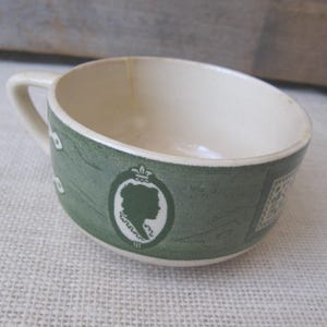 May include: A small, off-white teacup with a curved handle. The exterior features a green patterned design with a cameo silhouette and decorative elements. The interior is a creamy white color.