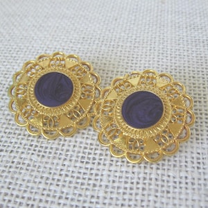 May include: A pair of gold-toned, filigree-style earrings with a circular, deep purple enamel center. The earrings have a scalloped edge and are set against a neutral background.