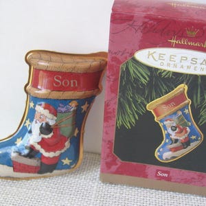 May include: A Hallmark Keepsake Ornament in the shape of a blue Christmas stocking with a red cuff labeled Son. The stocking features a graphic of Santa Claus standing on a chimney with a green sack. The ornament comes in a red Hallmark box.