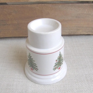 May include: White ceramic candle holder with a red trim and Christmas tree design. The candle holder has a wide base and a cylindrical top, perfect for holding a small candle. The Christmas tree design features green trees with red ornaments.