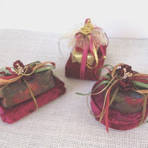 May include: Three decorative gift boxes wrapped in dark velvet fabric. Each box is adorned with gold cord, multi-colored ribbons in shades of red, green, and gold, and small artificial floral accents. The boxes are rectangular, square, and round.